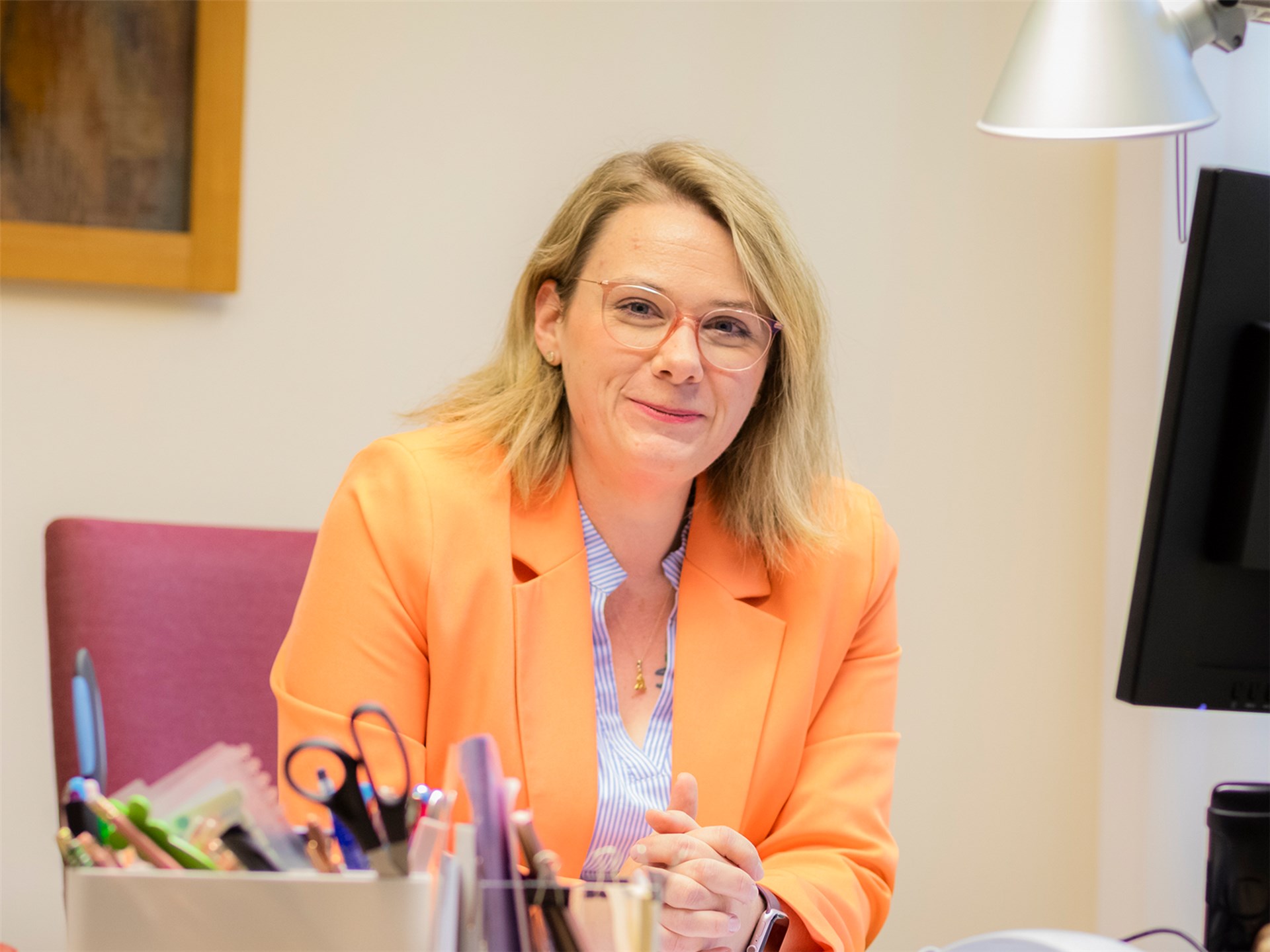 Frau mit orangefarbenen Blazer und Brille sitzt am Schreibtisch und lächelt in die Kamera